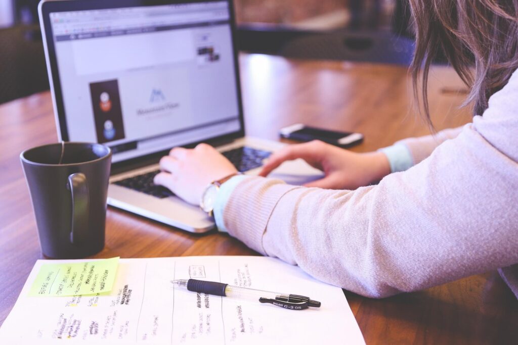 صورة ليد فتاة تكتب على اللابتوب مع دفتر وكوب قهوة